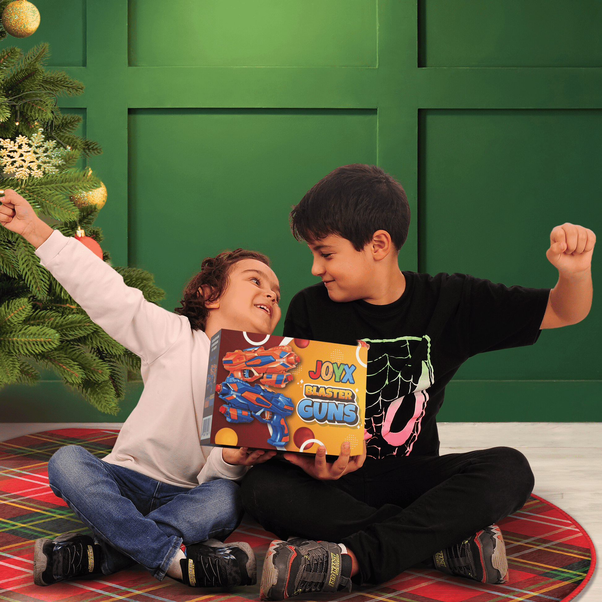 Two joyful children sitting by a Christmas tree, excitedly holding a box of JoyX Blaster Guns, celebrating a festive gift moment.