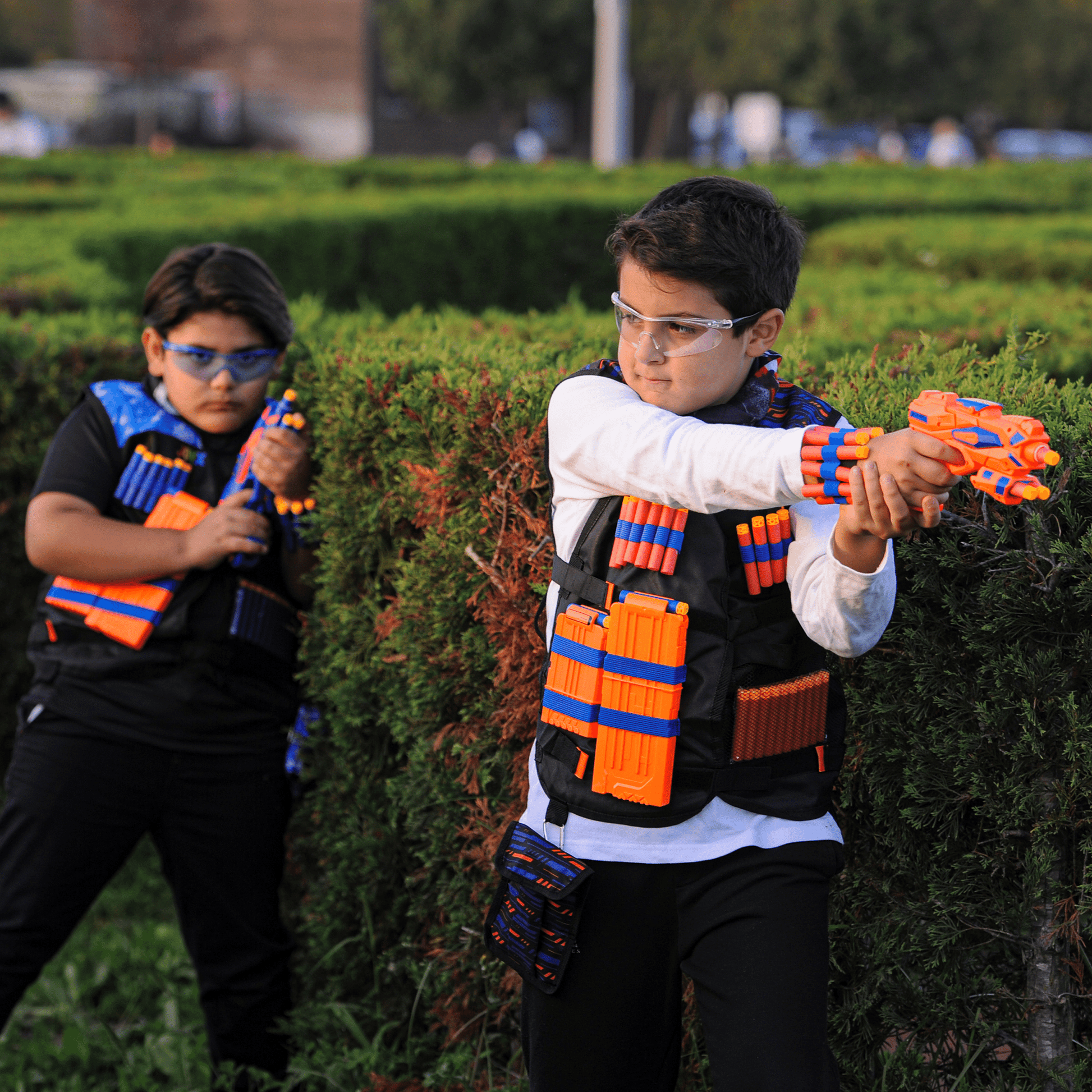 Two kids playing outdoors with JoyX blaster guns and wearing tactical vests filled with foam darts.